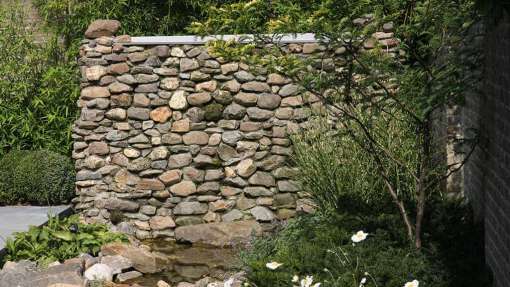 watermuur met opslagtank van 2500 liter water zodat er voldoende buffer aanwezig is. op de voorgrond een meerstammige solitaier struik gleditsia triacanthus hoogte muur 170 cm en breed 325 cm met maaskeien